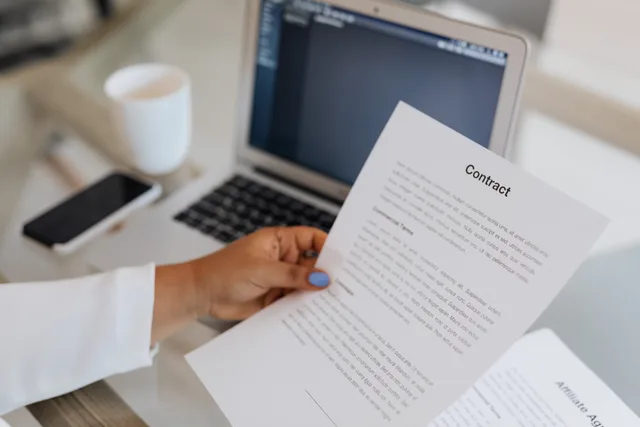 Close-up of a person holding a contract document next to a laptop during a work session. Mäta byråns effekt: KPI:er, rapportering och utvärdering i avtal